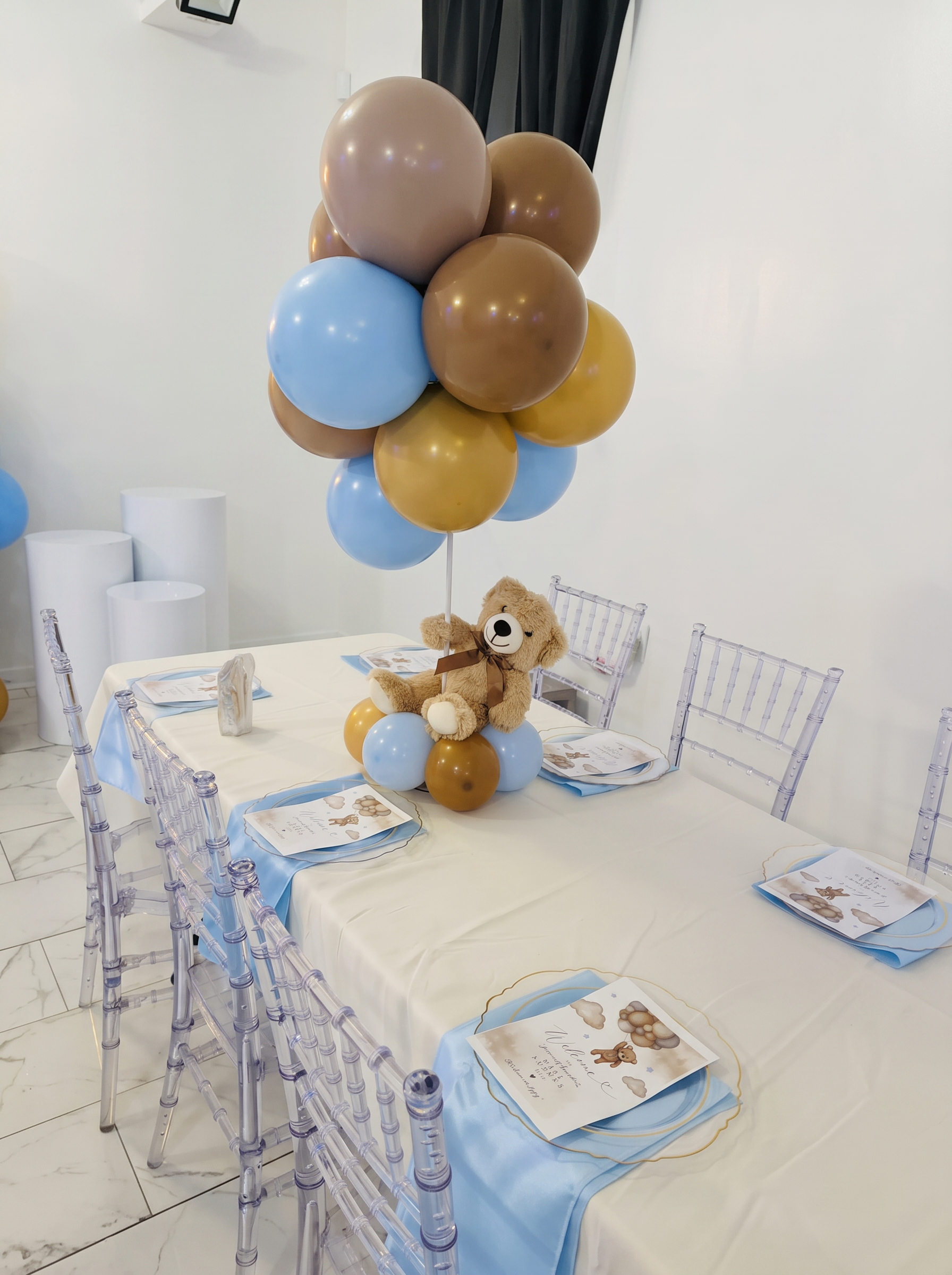 Baby shower guest table with a teddy bear balloon centerpiece, blue satin runner and clear chiavari chairs