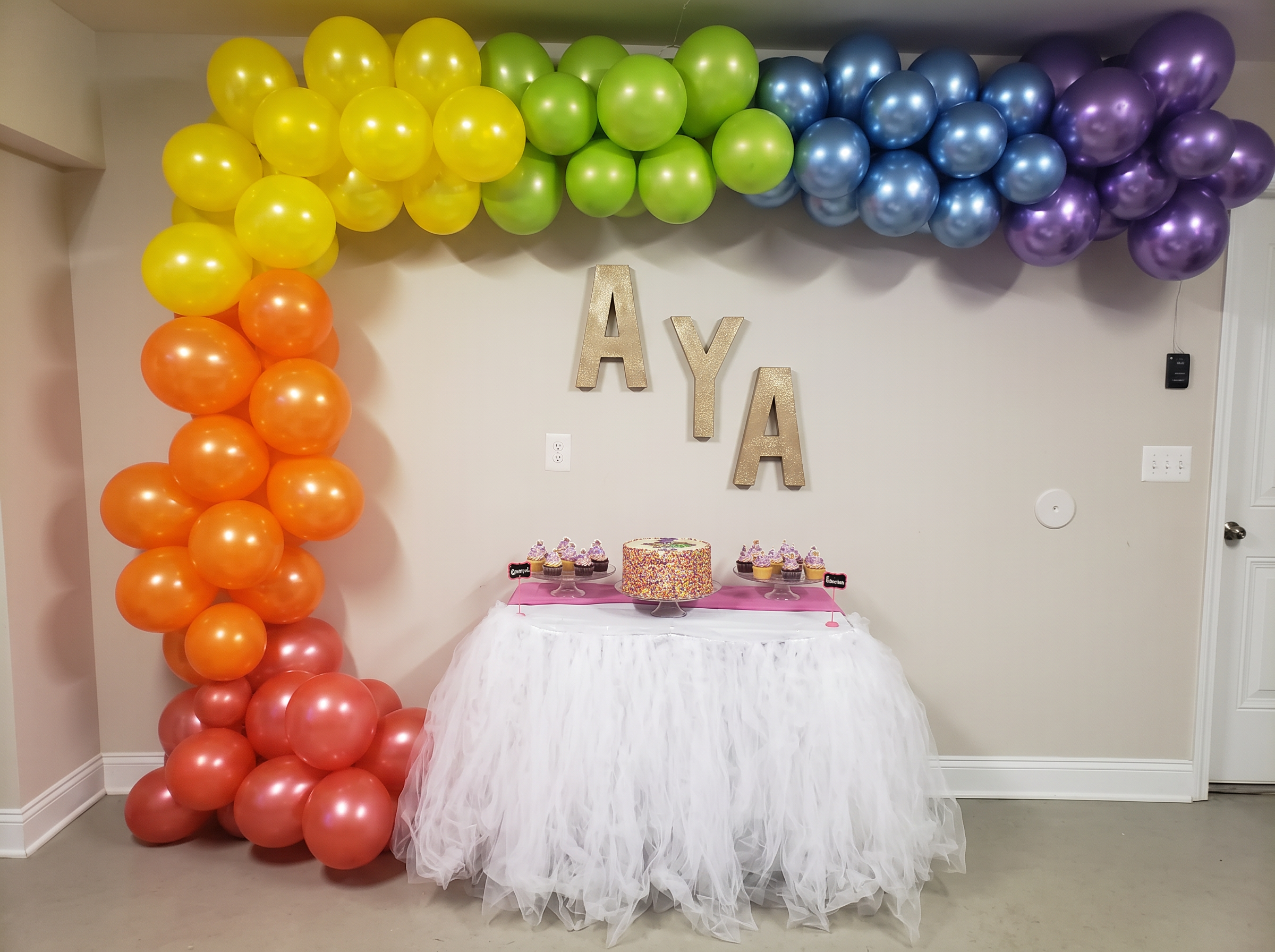 First birthday rainbow balloon arch framing a tulle-skirted cake table with gold name letters