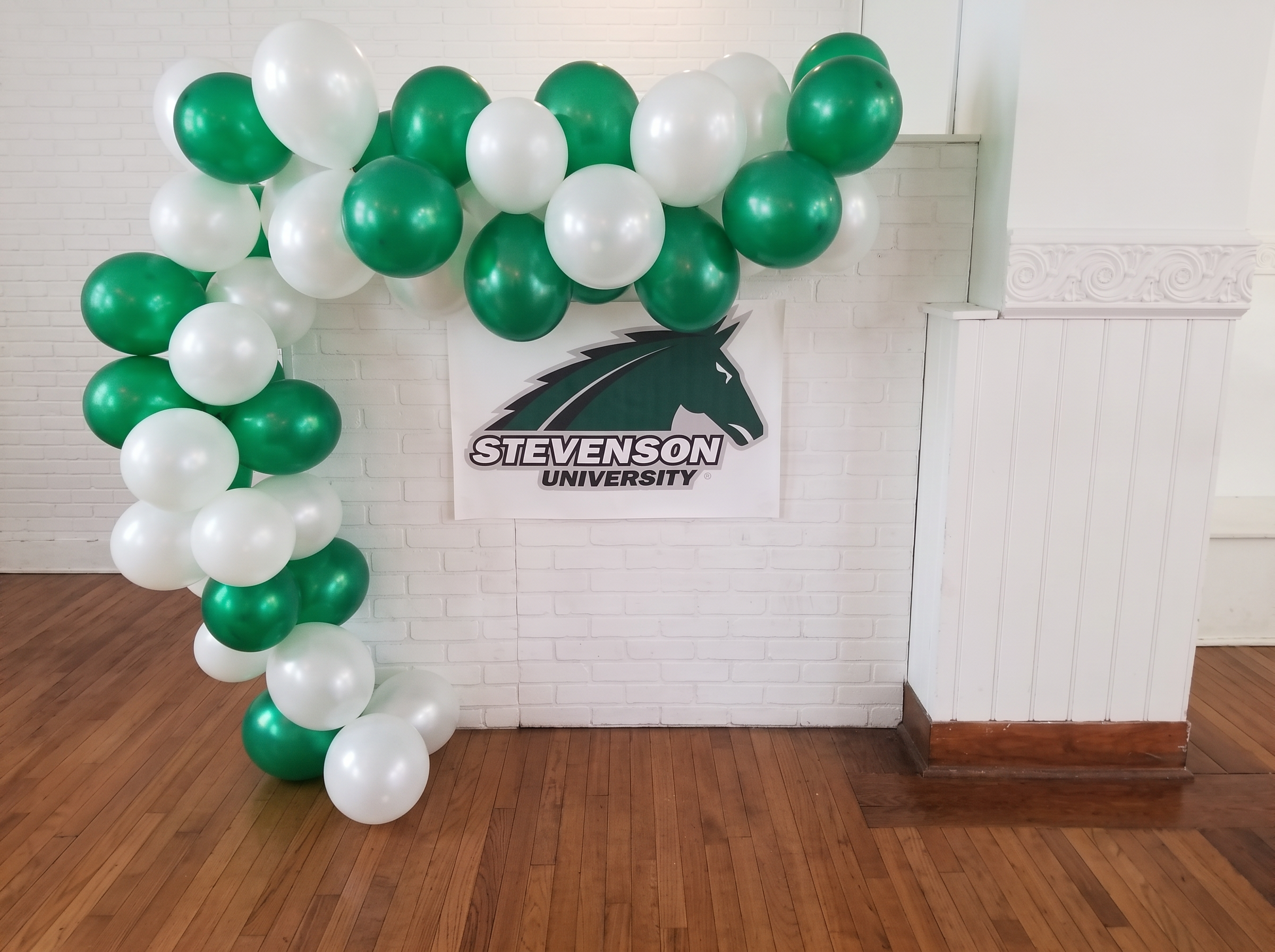 Green and white balloon arch around a university banner on a white brick wall