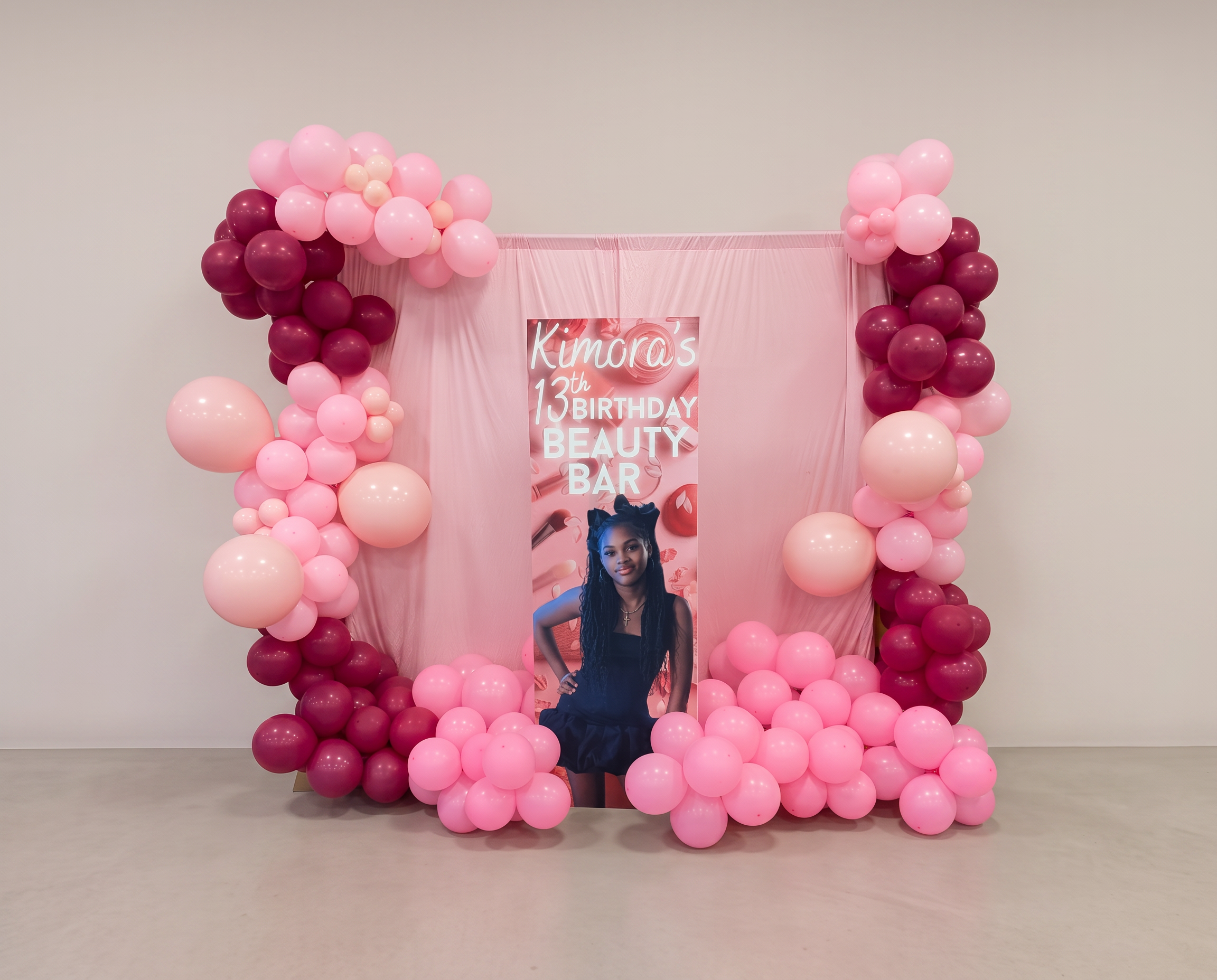 Pink and burgundy balloon frame around a pink draped backdrop with a custom beauty bar banner
