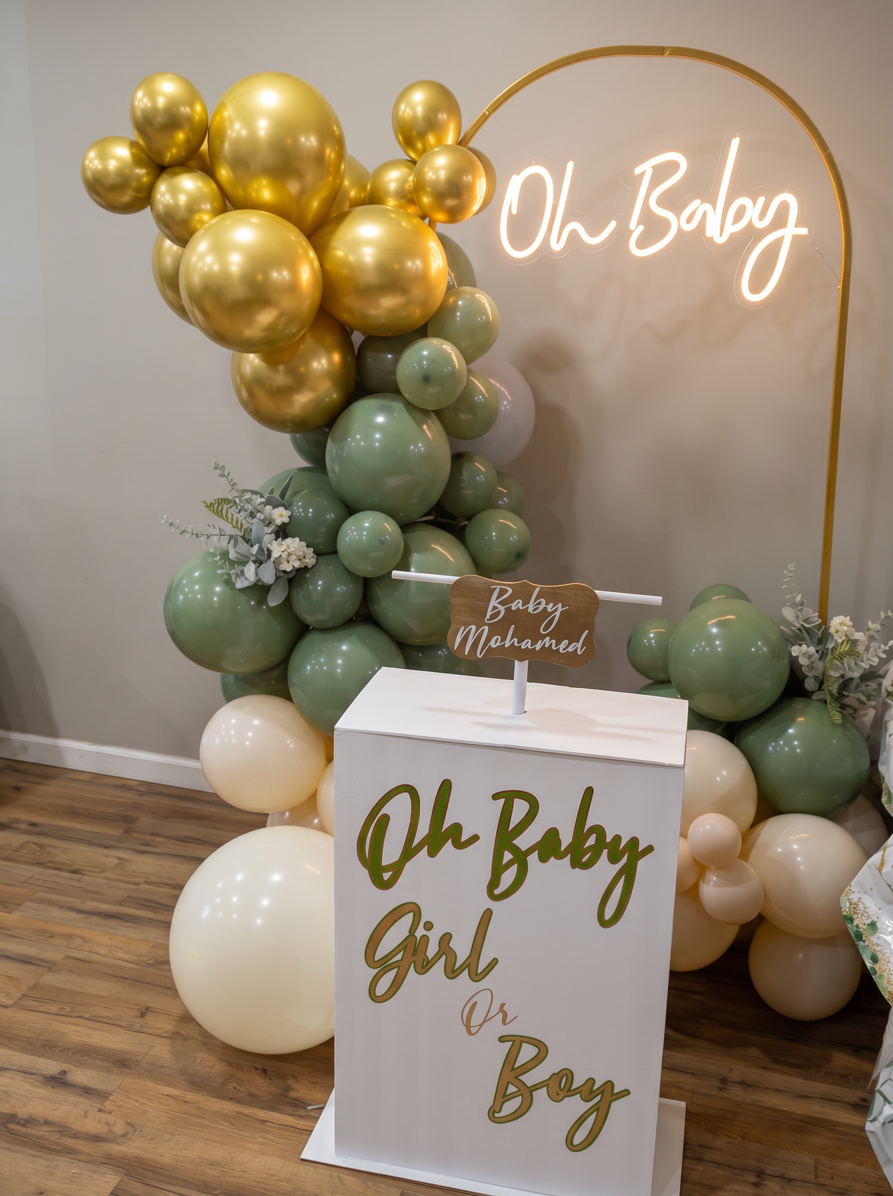 Gender reveal setup with a sage and gold balloon garland, Oh Baby neon sign and a lettered white plinth