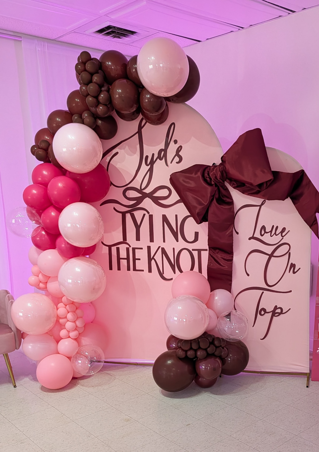 Pink double arch backdrop with a large burgundy bow and a Tying the Knot sign