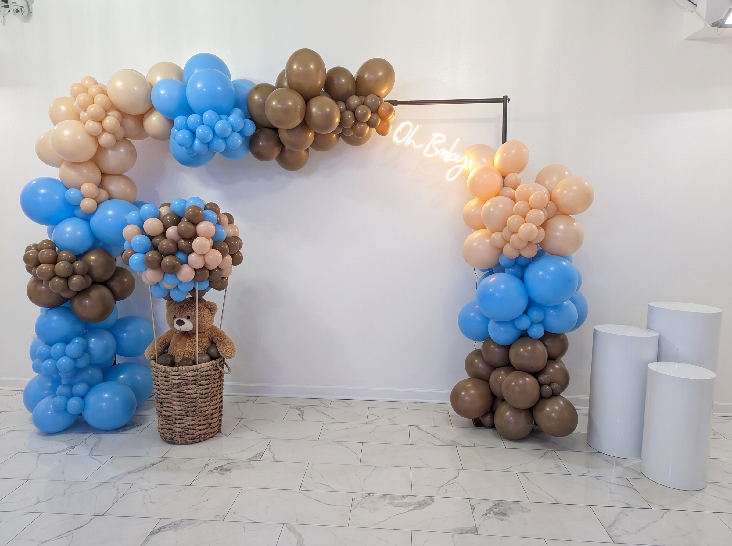 Blue, taupe and blush balloon arch with an Oh Baby neon sign, teddy bear balloon basket and white cylinder plinths