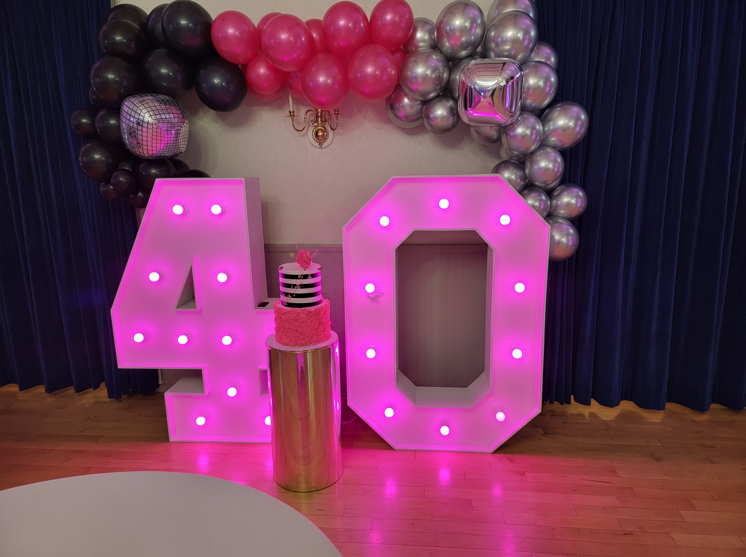 Lighted marquee 40 numbers with a pink, black and silver balloon garland and a striped cake on a gold plinth