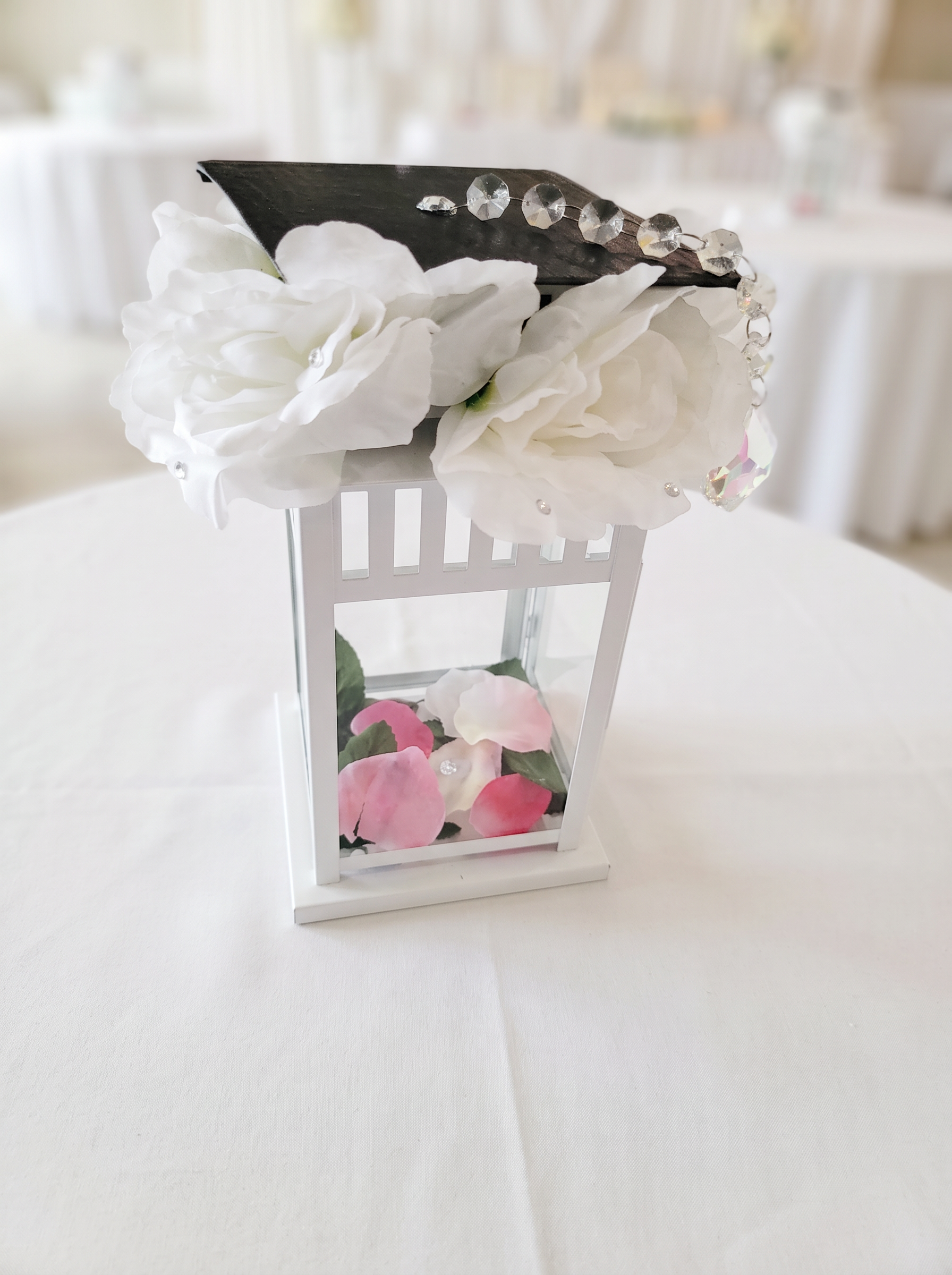 Graduation lantern centerpiece topped with a cap, white roses, crystals and pink rose petals inside
