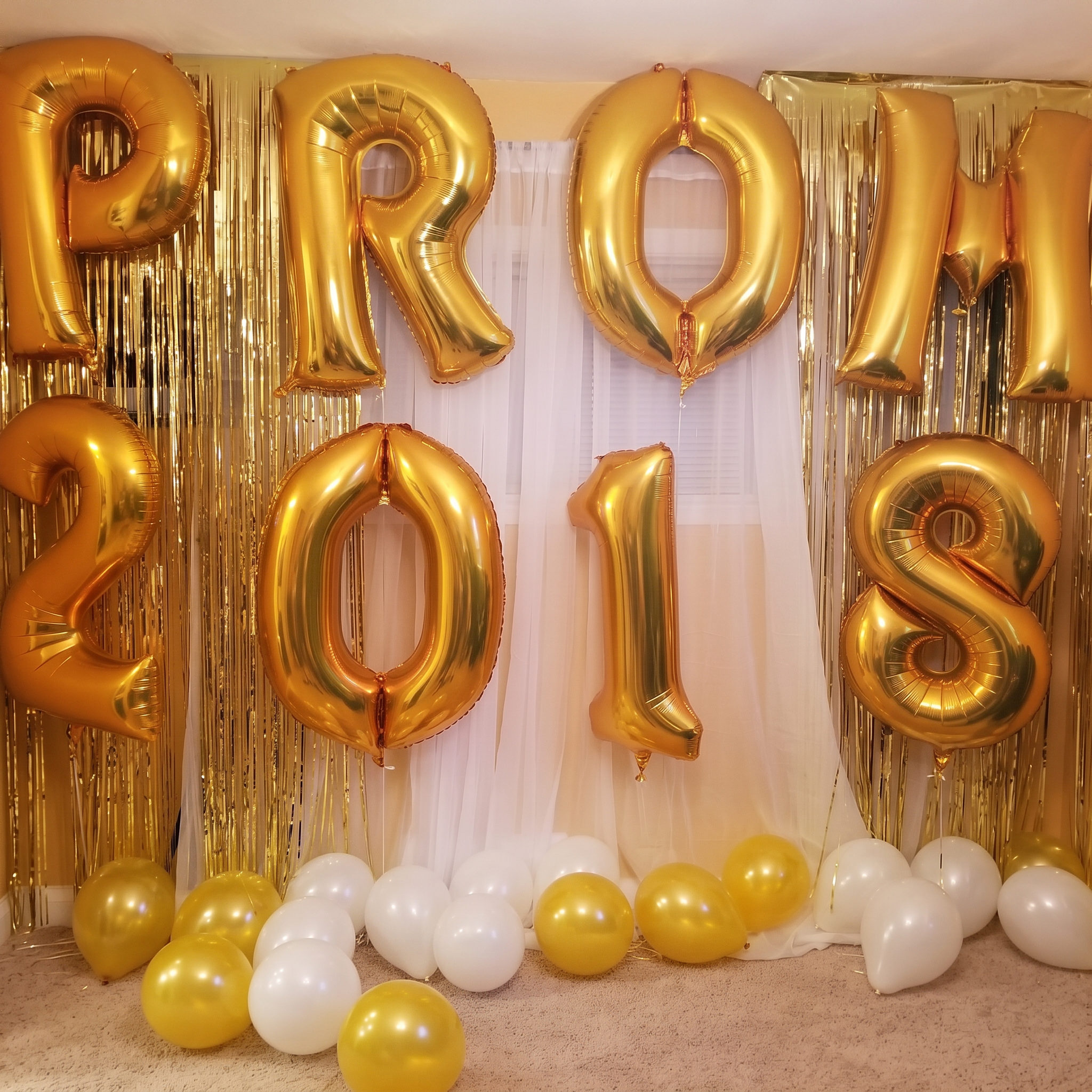 Gold foil balloon letters spelling prom above a gold fringe and white draping backdrop