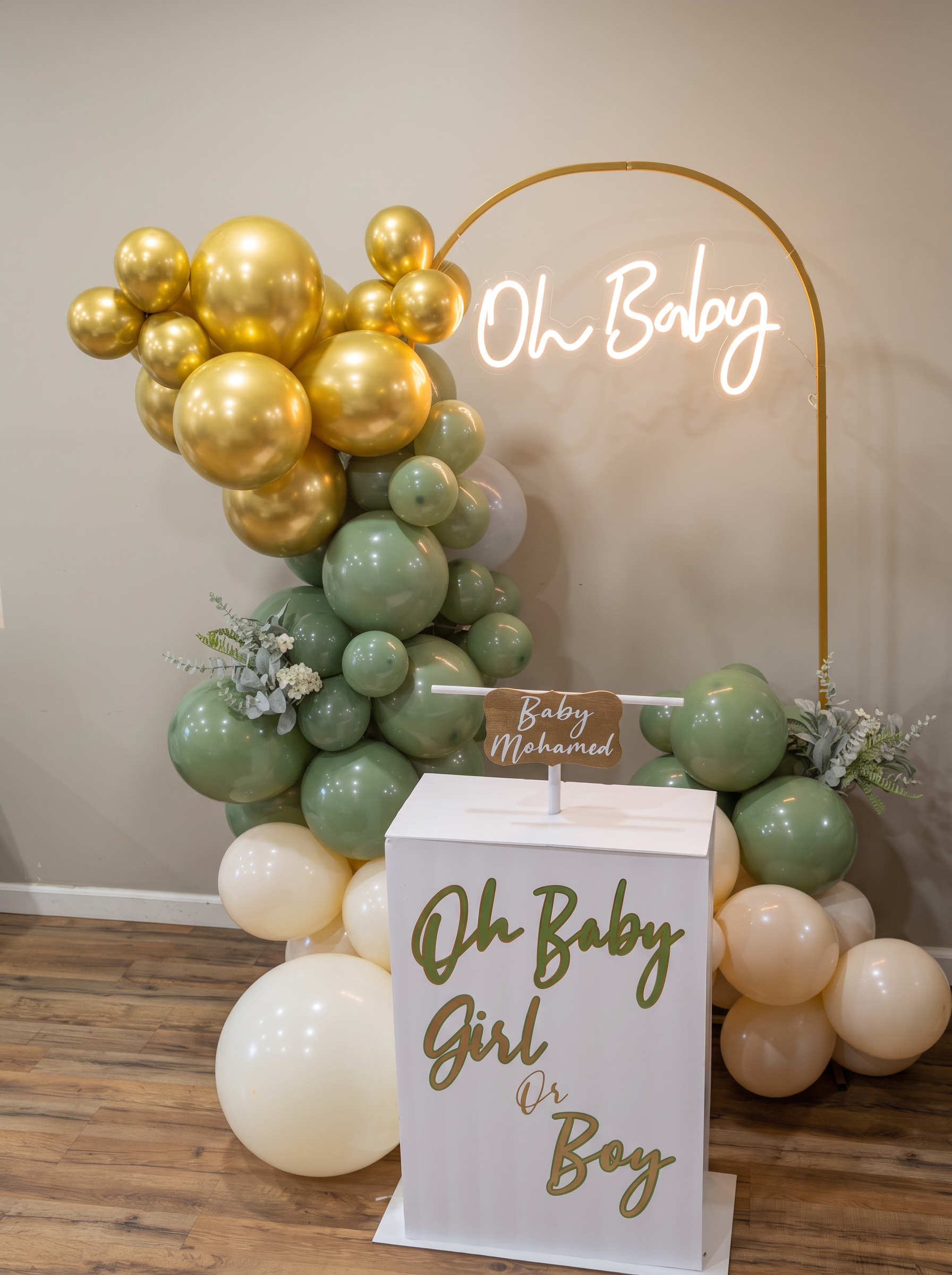Gold arch stand holding an Oh Baby neon sign beside a sage and gold balloon garland and eucalyptus florals
